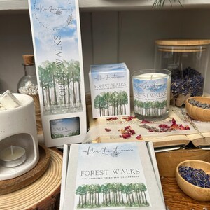 May include: A selection of scented candles and wax melts from The New Forest Candle Co. The products are labelled "Forest Walks" and feature a forest design. The image includes a tall box, a square box, and a glass candle.