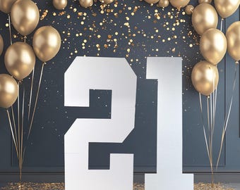 May include: A large white number 21 standing upright, surrounded by metallic gold balloons and scattered gold confetti against a dark blue background. The number 21 is displayed in a bold, block font, creating a festive display for a celebration.