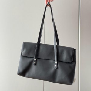 Può includere: Una borsa tote in pelle nera con dettagli in argento. La borsa ha una chiusura a lembo e due manici superiori.