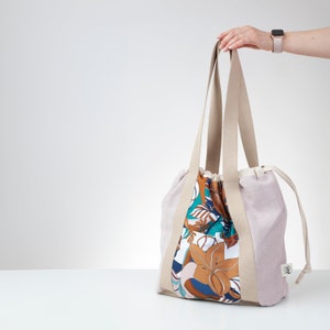 Op de afbeelding: Een beige tote bag met een trekkoord sluiting en lange hengsels. De tas is gemaakt van linnen stof en heeft een bloemenprint voering in tinten bruin, teal en wit.
