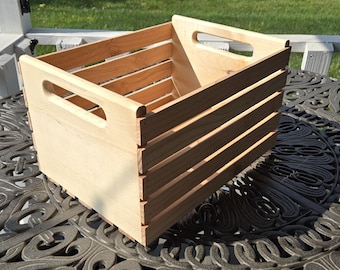Peut inclure: Une caisse en bois de couleur claire, de forme rectangulaire, avec des poignées découpées. La caisse comporte des lattes horizontales sur les côtés et repose sur une table en métal décorative. L'arrière-plan comprend une balustrade de porche blanche et de l'herbe verte.