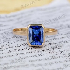 Może przedstawiać: Złoty pierścionek z dużym, kwadratowym, niebieskim szafirem. Pierścionek leży na białym tle.