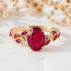 Peut inclure: Une bague en or ornée d'un grand rubis ovale en pierre centrale. La monture est ornée de petites pierres rouges et de détails délicats en forme de feuilles, avec de petits diamants. La bague est posée sur un fond doux et neutre.