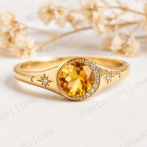 Könnte beinhalten: Ein goldener Ring mit einem großen, runden, gelben Edelstein. Der Ring hat ein himmlisches Design mit einem Halbmond, Sternen und kleinen Diamanten. Der Ring liegt auf einer weißen Oberfläche mit getrockneten Blumen im Hintergrund.