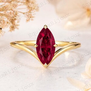 Puede incluir: Un anillo de oro con una piedra preciosa de color rojo intenso en talla marquesa montada en una banda en forma de V. El metal tiene un acabado pulido y la piedra está asegurada por cuatro garras. El diseño crea una silueta puntiaguda.