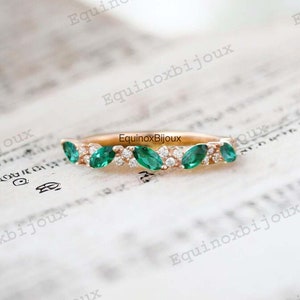 May include: A rose gold ring featuring marquise-cut emeralds and small round diamonds. The emeralds are set in a row, alternating with the diamonds. The ring is on a textured, off-white surface. The brand name "EquinoxBijoux" is visible.