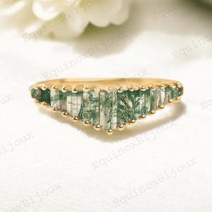 May include: A gold ring featuring a series of green and white rectangular gemstones arranged in a V-shape. The ring is displayed on a white surface, with a blurred white flower in the background. A piece of jewellery.