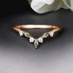 May include: A rose gold ring with a curved design. The ring features small, round, clear stones and marquise-shaped green stones. The ring is set against a dark background with white flowers in the background.