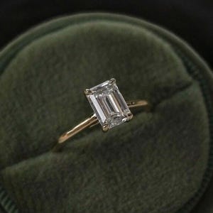 May include: A gold ring featuring a large, rectangular, clear gemstone. The ring is set in a gold band and is presented in a dark green velvet ring box. The gemstone has a clear, faceted appearance.