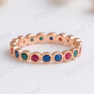 May include: Rose gold eternity ring with alternating red, green, and blue gemstones. The ring features a delicate design with a series of bezel-set stones. The gemstones are round and evenly spaced around the band.