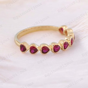 Puede incluir: Un anillo de oro con una hilera de gemas rojas en forma de corazón. El anillo tiene una banda de oro pulido y las gemas están espaciadas uniformemente. El anillo se muestra sobre un fondo suave y blanquecino.