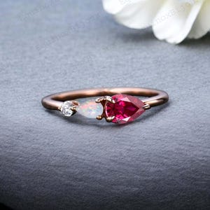 May include: A rose gold ring featuring three gemstones. A small, round, clear gemstone is set on one side, with a pear-shaped opal and a larger, pear-shaped, deep red gemstone in the center. The ring is on a gray surface.