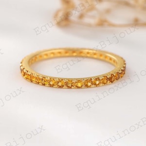 Op de afbeelding: Een goudkleurige ring met een band van kleine, gele edelstenen. De ring staat tegen een zachte, witte achtergrond. De ring is een delicaat sieraad.