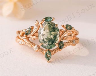 May include: A rose gold engagement ring set with a large oval green moss agate stone and smaller green accent stones. The band has a nature-inspired design with leaf and branch details, and small white accent stones.
