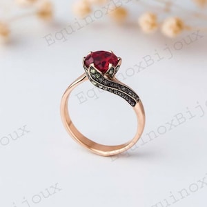 Op de afbeelding: Roségouden ring met een opvallende rode edelsteen. De band heeft een vloeiend ontwerp, versierd met donkere en groene stenen. Het ontwerp van de ring is geplaatst tegen een zachte, neutrale achtergrond.
