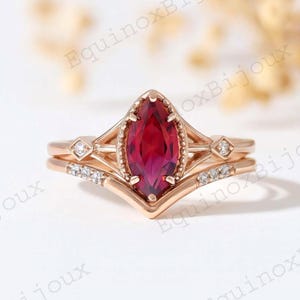 Könnte beinhalten: Ein roségoldenes Ring-Set mit einem markant geschliffenen, tiefroten Edelstein im Zentrum. Der Verlobungsring wird von zwei Bändern flankiert, eines mit kleinen Diamanten und das andere mit einem Diamant-Akzent. Das Set hat ein Vintage- und elegantes Design.