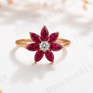 May include: A gold ring featuring a flower design. The flower has deep red petals surrounding a central, round, clear gemstone. The ring is set against a soft, white background with delicate floral accents.