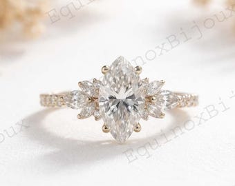 Peut inclure: Une bague de fiançailles en or avec une grosse pierre centrale en forme de marquise et de plus petites pierres en forme de pétales l'entourant. La bande est également ornée de petites pierres scintillantes. La bague est posée sur un fond flou et doux.
