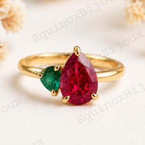 Könnte beinhalten: Ein goldener Ring mit einem birnenförmigen, tiefroten Edelstein und einem kleineren, herzförmigen grünen Edelstein. Der Ring hat ein schlichtes Band und liegt auf einem neutralen Hintergrund.