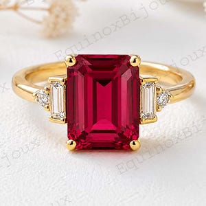 May include: A gold ring featuring a large emerald-cut red gemstone in the centre, flanked by two baguette-cut clear stones and two small round clear stones on each side, set on a polished yellow gold band with a smooth, rounded finish.
