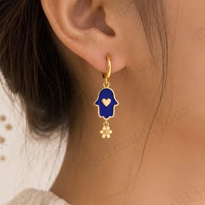 May include: Gold-coloured hoop earring featuring a blue Hamsa hand charm and a small gold flower pendant. The Hamsa hand has a gold heart in the centre. The earring is worn on a person's ear.