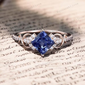 May include: A rose gold engagement ring featuring a square-shaped blue sapphire centre stone surrounded by smaller diamonds. The ring has a delicate, intricate design with intertwined bands.