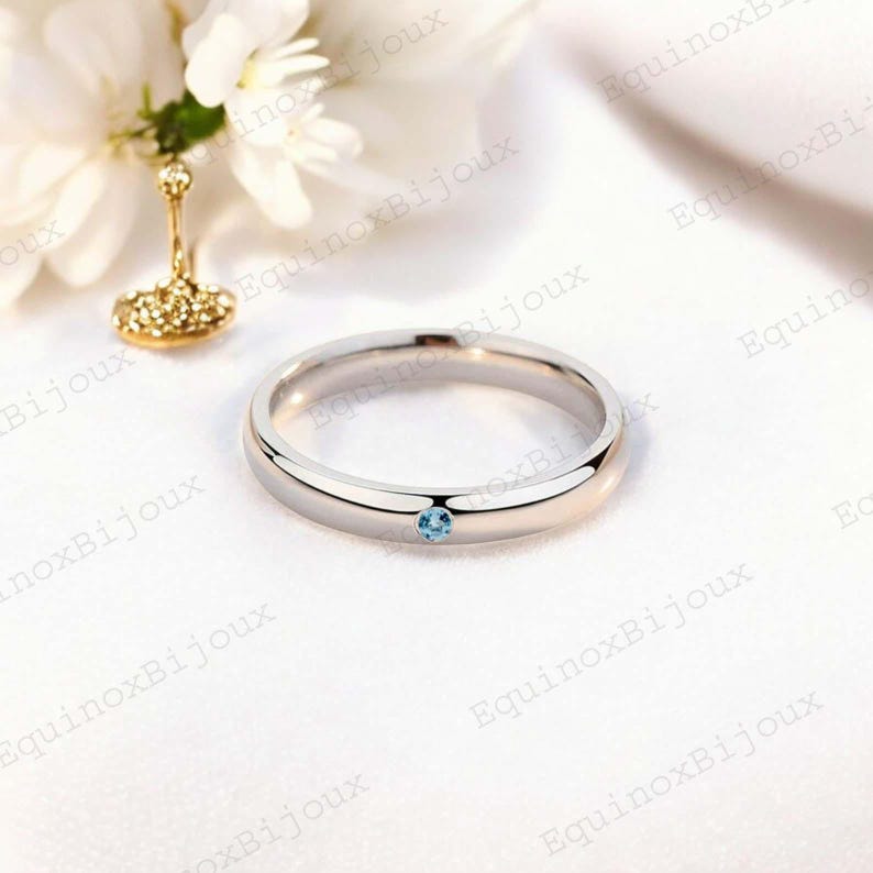 May include: A silver ring with a small, round, light blue gemstone set in the center. The ring is smooth and polished, reflecting the light. The background is a soft white, with a blurred floral arrangement and gold jewelry in the background.