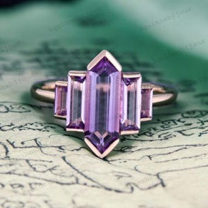 May include: A rose gold ring featuring a large, rectangular, purple amethyst gemstone in the centre. The amethyst is surrounded by three smaller, rectangular, purple amethyst gemstones.