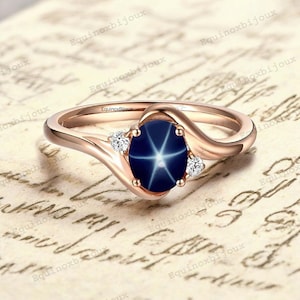 Peut inclure: Une bague en or rose avec une pierre précieuse saphir bleu étoilé et deux diamants plus petits. La bague a une bande incurvée.