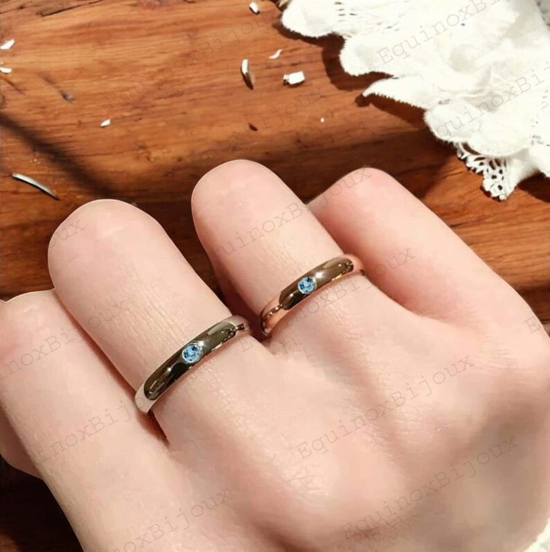 May include: Two rings, one silver and one gold, each featuring a small, round, light blue gemstone. The rings are displayed on fingers, showcasing their simple band design and the subtle sparkle of the gemstones. The background is a warm, wooden surface.