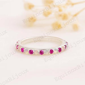 May include: A silver ring with a band of alternating pink and white gemstones. The ring is set against a soft, blurred background. The ring is a piece of jewelry.