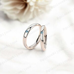 May include: Two wedding rings with a rose gold band and a white inlay. Each ring features a small, round, light blue gemstone. The rings are displayed on a white surface, with a blurred floral background.