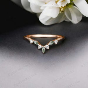 May include: A delicate rose gold wedding band with a V-shaped design featuring small, clear and green gemstones.