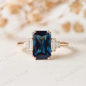 May include: Rose gold ring featuring a large, rectangular, deep blue gemstone as the centerpiece. The ring is flanked by smaller, rectangular, clear gemstones on each side. The band is a delicate rose gold color, and the overall design is elegant.