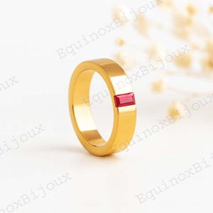 May include: A gold-colored ring with a rectangular, red gemstone. The ring has a flat band and a polished finish. The gemstone is set into the ring's design, adding a pop of color. The ring is displayed on a white surface.