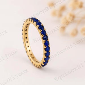 以下が含まれることがあります： 小さな丸い濃い青色の宝石が並んだ、金色のリング。リングは柔らかくぼやけた背景に配置されています。宝石はバンド全体に均等に配置されています。