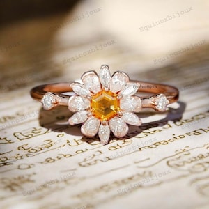 Può includere: Un anello in oro rosa con una grande pietra preziosa gialla circondata da pietre bianche più piccole, incastonato in un design a forma di margherita. L'anello è su una superficie testurizzata di colore crema.