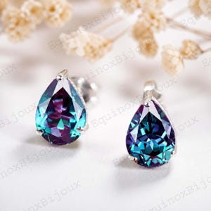 May include: A pair of teardrop-shaped earrings with a silver setting. The gemstones have a color-shifting effect, displaying shades of teal, purple, and blue. The earrings are set against a soft, white background with blurred floral accents.