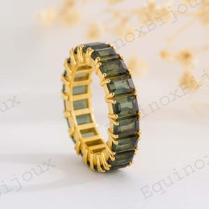 May include: A gold-colored ring with a band of rectangular, dark green gemstones. The ring is set against a soft, blurred background, highlighting the intricate details of the jewelry. The gemstones are evenly spaced around the band.