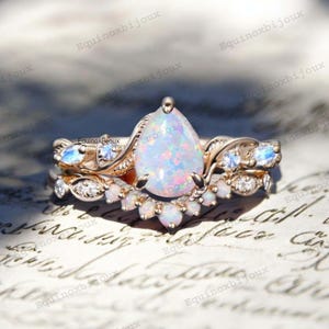 May include: A rose gold ring set with a pear-shaped opal centre stone and a matching band. The opal displays a vibrant play of colour. The bands are adorned with smaller opals and accent stones. The text "Equinoxbijoux" is visible.