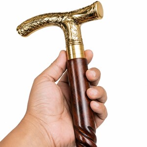 Puede incluir: Un bast&oacute;n con un mango en forma de T de metal dorado, decorado con intrincados patrones florales en relieve. El mango se une a un eje de madera de color marr&oacute;n oscuro que presenta una textura tallada en espiral en la parte superior.