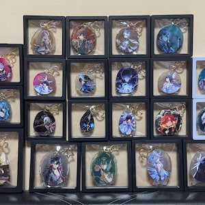 Puede incluir: Colección de llaveros de personajes exhibidos en pequeñas cajas de sombra en blanco y negro. Cada llavero presenta una ilustración de personaje de estilo anime diferente con herrajes dorados. Los llaveros son de varios colores, incluyendo rojo, azul y morado.