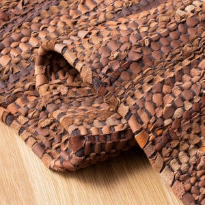 May include: A close-up of a brown leather rug with a woven design. The rug features various shades of brown, creating a textured and layered appearance. The edges of the rug are slightly curled, revealing the underside. The rug is placed on a light-colored wooden surface.