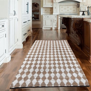 May include: A white and brown diamond patterned rug with a black border. The rug is laid out on a hardwood floor in a kitchen.