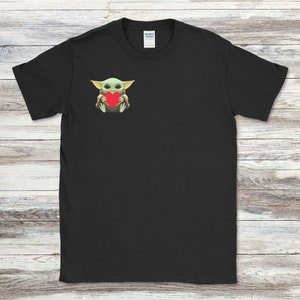 May include: Black t-shirt featuring a graphic of a green alien-like character holding a red heart. The character has large eyes and pointed ears. The t-shirt is made of a soft material.