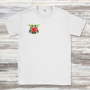 May include: White t-shirt featuring a graphic of a green alien-like character holding a red heart. The character has large eyes and pointed ears. The shirt is made of a soft material and has a crew neck.