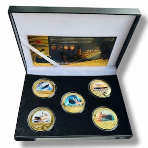 May include: A black velvet box with gold trim, containing a set of six gold-coloured coins featuring images of the RMS Titanic. The coins are arranged in a circular pattern, with the words "The Ship Majestic" and "RMS Titanic" printed on each coin. The box also includes a card with information about the Titanic.