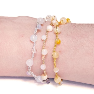 Peut inclure: Trois bracelets superposés avec des perles blanches, roses et jaunes. Les bracelets sont en chaîne dorée et argentée.