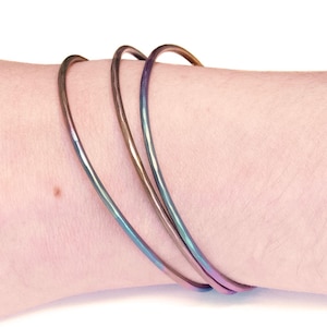Op de afbeelding: Drie iriserende metalen armbanden gestapeld op een pols. De armbanden zijn dun en hebben een gladde, gepolijste afwerking. De kleuren veranderen van blauw naar paars naar roze.