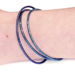 Peut inclure: Trois bracelets fins en métal empilés sur un poignet. Les bracelets sont un dégradé de bleu et de violet.